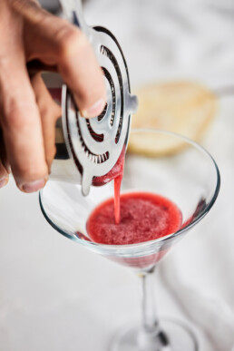 Pouring a mix of fresh juices through a metal sieve into a martini glass